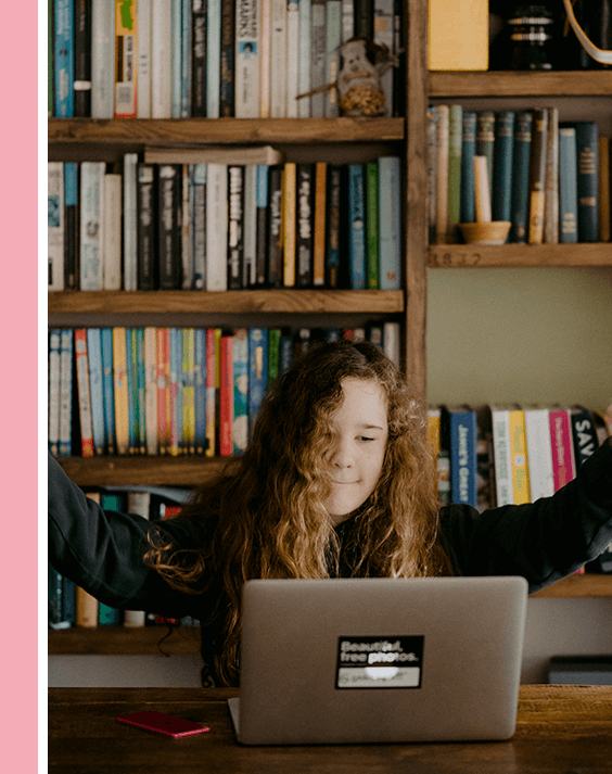 A girl sitting at a table with her arms raised in front of a laptop.